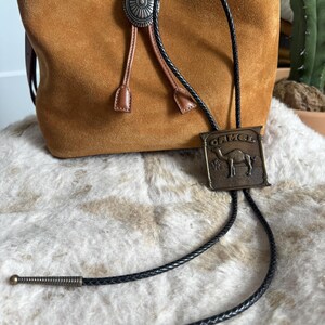 May include: A brown suede drawstring bag with a silver concho and a black bolo tie featuring a rectangular metal plate with a camel image and the word "CAMEL". The tie has decorative metal tips.