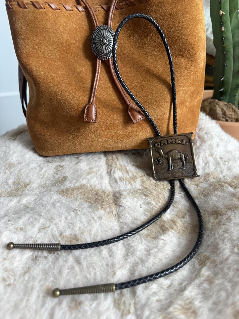 May include: A brown suede drawstring bag with a silver concho and a black bolo tie featuring a rectangular metal plate with a camel image and the word "CAMEL". The tie has decorative metal tips.