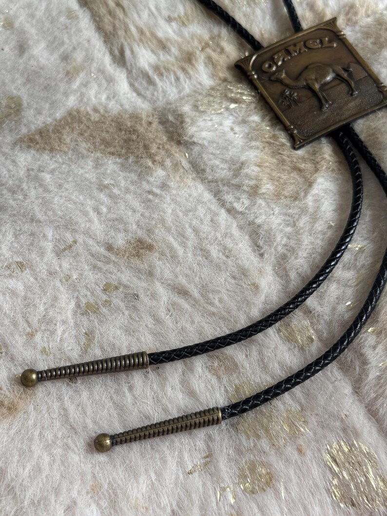 May include: A bolo tie featuring a rectangular bronze-colored pendant with a camel illustration and the word "CAMEL". The tie has a black braided cord with decorative metal tips.