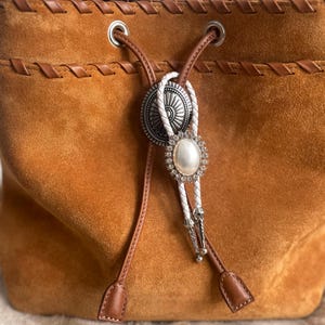 May include: A suede bucket bag in a warm brown hue, featuring a drawstring closure and decorative stitching. The bag is accented with a bolo tie featuring a silver medallion and a large pearl-like embellishment. The bag's leather details add a touch of rustic elegance.