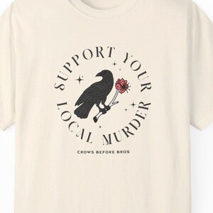 Peut inclure: Un t-shirt à manches courtes de couleur crème avec un motif noir représentant un corbeau perché sur une branche tenant une fleur rouge. Le texte SUPPORT YOUR LOCAL MURDER est écrit en cercle autour de l'oiseau, avec CROWS BEFORE BROS en dessous.