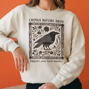 Pode incluir: Uma camisola de gola redonda cor de creme com uma impressão gráfica preta de um corvo rodeado por elementos florais dentro de uma borda decorativa. O texto "CROWS BEFORE BROS" aparece acima e "Support your local murder." abaixo.