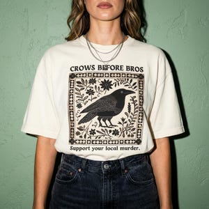 May include: An off-white short-sleeve t-shirt featuring a black graphic of a crow surrounded by floral patterns within a square border. The text reads Crows Before Bros and Support your local murder. The shirt is paired with dark denim jeans.