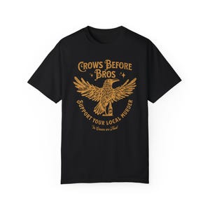 Peut inclure: Un t-shirt noir à manches courtes avec un motif doré représentant un corbeau perché sur une bouteille. Le texte indique : CROWS BEFORE BROS, SUPPORT YOUR LOCAL MURDER et In Crows we Trust. Le design inclut des étoiles et une texture usée.