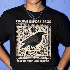 Puede incluir: Una camiseta negra de manga corta con un gráfico cuadrado de color crema que muestra una ilustración de un cuervo negro rodeado de motivos florales. El texto "CROWS BEFORE BROS" aparece arriba y "Support your local murder." abajo.