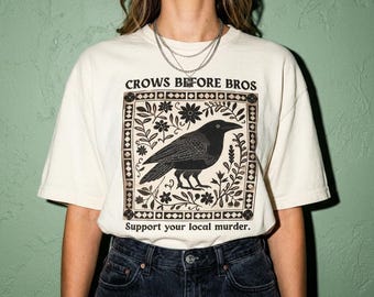 May include: An off-white short-sleeve t-shirt featuring a black graphic of a crow surrounded by floral patterns within a square border. The text reads "CROWS BEFORE BROS" at the top and "Support your local murder." at the bottom in black lettering.