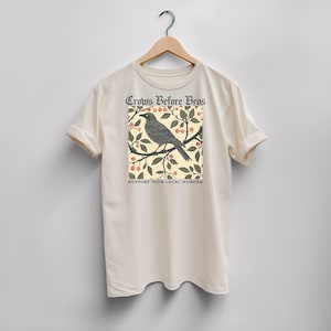 Peut inclure: Un t-shirt à manches courtes de couleur crème avec un motif représentant un corbeau noir perché sur une branche avec des feuilles et des baies. Le texte "Crows Before Bros" figure au-dessus et "SUPPORT YOUR LOCAL MURDER" en dessous.