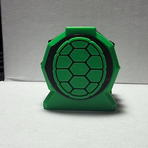 May include: A 3D-printed green and black object with a geometric design. The object has a rounded top with a hexagonal pattern in the center, surrounded by a black border. The base is a solid green color.