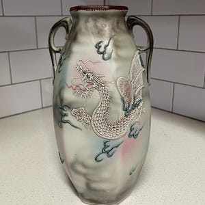 May include: A decorative vase with a dragon motif. The vase is a light gray with a dragon design in white and pink. It has two handles and a dark red rim. The dragon is surrounded by stylized clouds.