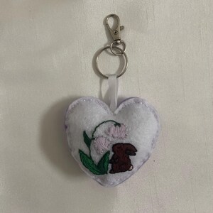 Pu&ograve; includere: Un portachiavi a forma di cuore in feltro bianco con un piccolo coniglio ricamato e fiori. Il portachiavi ha un anello di metallo argentato e un nastro bianco.