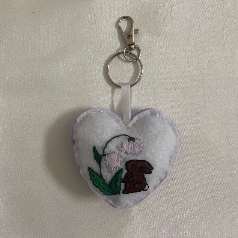 Pu&ograve; includere: Un portachiavi a forma di cuore in feltro bianco con un piccolo coniglio ricamato e fiori. Il portachiavi ha un anello di metallo argentato e un nastro bianco.