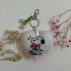 Pu&ograve; includere: Portachiavi a forma di cuore in feltro bianco con cuciture rosa. Il portachiavi presenta un'immagine ricamata di Snoopy che tiene un mazzo di rose rosse. Il portachiavi ha un anello in metallo argentato e un ciondolo con perline di fiori rosa e verdi.
