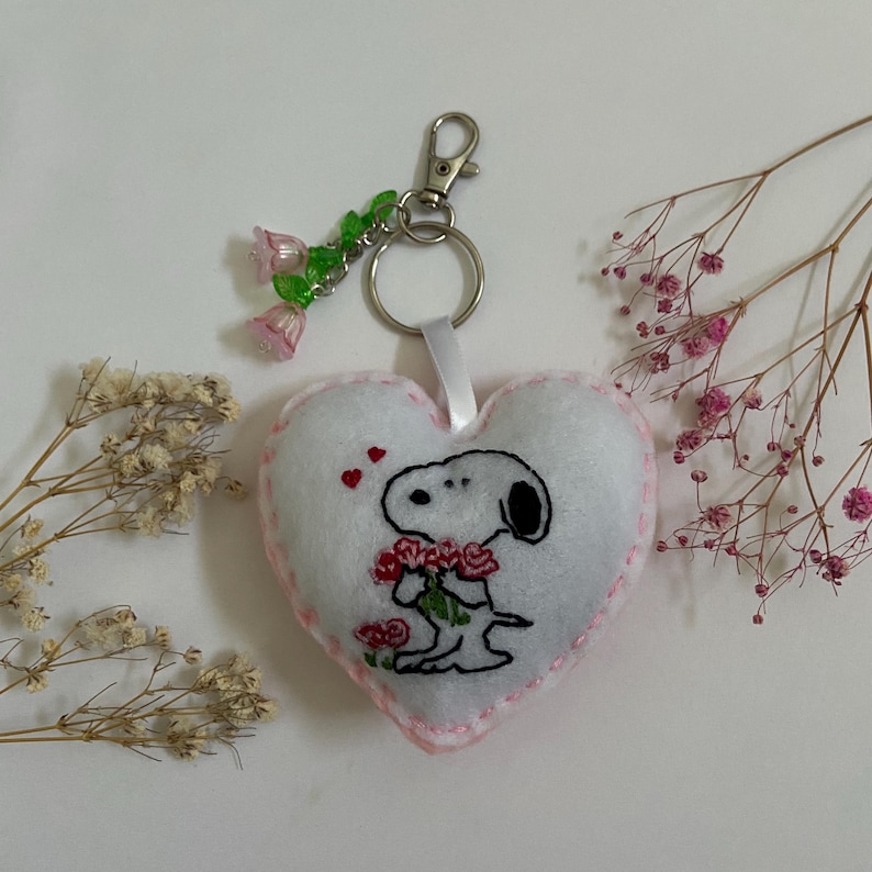 Pu&ograve; includere: Portachiavi a forma di cuore in feltro bianco con cuciture rosa. Il portachiavi presenta un'immagine ricamata di Snoopy che tiene un mazzo di rose rosse. Il portachiavi ha un anello in metallo argentato e un ciondolo con perline di fiori rosa e verdi.