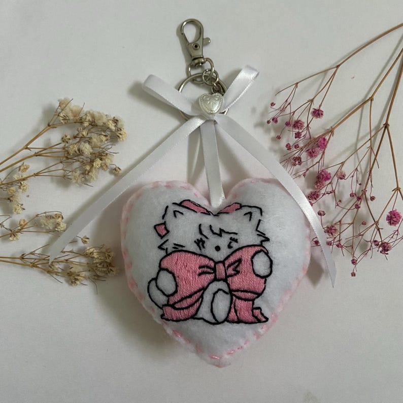 Pu&ograve; includere: Portachiavi a forma di cuore in feltro bianco con un gatto ricamato rosa che tiene un fiocco rosa. Il portachiavi ha un nastro bianco.