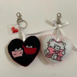 Pu&ograve; includere: Due portachiavi a forma di cuore in feltro. Un portachiavi &egrave; nero con un design a cartone animato rosso e rosa. L'altro portachiavi &egrave; bianco con un design a gatto di cartone animato rosa.