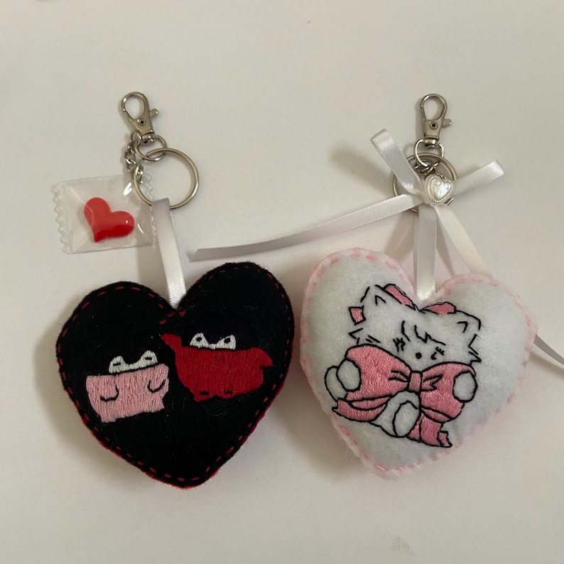 Pu&ograve; includere: Due portachiavi a forma di cuore in feltro. Un portachiavi &egrave; nero con un design a cartone animato rosso e rosa. L'altro portachiavi &egrave; bianco con un design a gatto di cartone animato rosa.