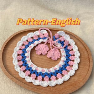May include: A crocheted collar necklace in white, pink, and blue with two pink crocheted flower embellishments. The necklace is on a wooden surface.