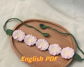 Puede incluir: Una diadema de crochet con flores rosas y amarillas y hojas verdes. La diadema está sobre un plato de madera. El texto "English PDF" está escrito en el plato.