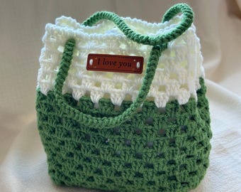 Puede incluir: Una bolsa de mano de crochet verde y blanca con una etiqueta de cuero marrón que dice "Te amo".