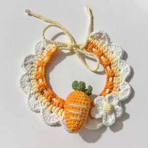 Könnte beinhalten: Ein gehäkeltes Halsband für Haustiere mit einer weißen und gelben Blume und einem Karotten-Anhänger. Das Halsband ist aus weißem und gelbem Garn gefertigt und hat einen gewellten Rand.