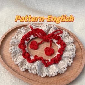 Peut inclure: Un collier crocheté avec un motif de cerise rouge. Le collier est blanc avec un volant rouge autour du bord. La cerise est faite de fil rouge et a une tige verte.