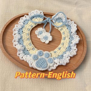 Puede incluir: Un collar de perro tejido a crochet con una huella de pata azul y blanca, dos flores blancas y un lazo amarillo y azul. El collar está sobre una superficie de madera. Pattern-English