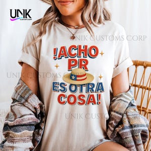 Op de afbeelding: Een crèmekleurig T-shirt met de tekst "!ACHO PR ES OTRA COSA!" in rood en blauw, met een strohoed met een Puerto Ricaans vlagontwerp. Het shirt heeft opgerolde mouwen.