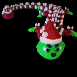Könnte beinhalten: Eine 3D-gedruckte Weihnachtsfigur einer grünen Kreatur mit rotem Weihnachtsmannhut und weißem Rand. Die Figur hat einen langen, zuckerstangen-gestreiften Schwanz und rote und weiße Akzente. Die Figur hat einen grimmigen Gesichtsausdruck.