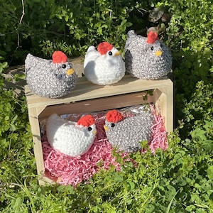 May include: Four crocheted chicken figurines, two white and two gray with red combs, displayed in a wooden crate with pink shredded paper.