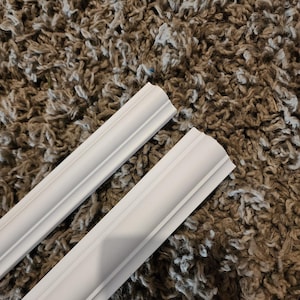 Puede incluir: Dos molduras decorativas blancas con un dise&ntilde;o curvo detallado. La moldura est&aacute; hecha de un material liso y se coloca sobre un fondo marr&oacute;n texturizado. Las piezas son probablemente para dise&ntilde;o de interiores o proyectos de mejoras para el hogar.