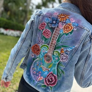 May include: A denim jacket with a hand-painted design of a guitar and flowers. The guitar is blue, green, and yellow, and the flowers are pink, red, orange, and purple.