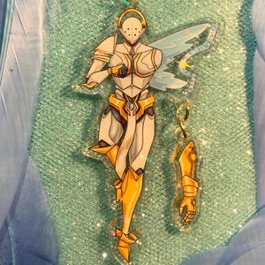 May include: A clear acrylic charm depicting a white and gold armoured figure, with a matching arm charm. The figure has blue wings and is attached to a gold star-shaped keychain. The background is a glittery teal.