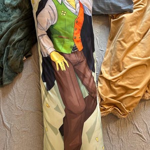 May include: A rectangular body pillow featuring a printed illustration of a smiling character with spiky yellow hair. The character wears a white collared shirt, a green and orange vest, brown trousers, and a dark jacket, set against a light background.