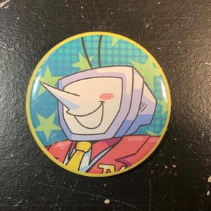 May include: A round button with a cartoon character design. The character has a white face, a TV-shaped head, and a long nose. The background is blue with green stars and dots, and the border is yellow.