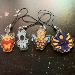 May include: Four clear acrylic charms with black lanyards. Each charm features a unique cartoon character design in vibrant colors: red and yellow, blue and white, gold and blue, and blue and yellow.