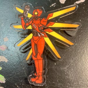 Może przedstawiać: Akrylowy brelok przedstawiający robota trzymającego pistolet. Robot ma dużą głowę z wizjerem i czerwone ciało z żółtymi akcentami. Tło to żółte promienie słońca.