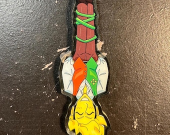 May include: An acrylic keychain featuring an upside-down cartoon character with spiky yellow hair, wearing a white shirt with an orange and green vest. The character's legs are bound by green vines, and the figure is attached to a lime green ring.