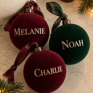 May include: Three round velvet Christmas ornaments in burgundy and dark green. Each ornament features a gold metallic cap, a ribbon tie, and a name printed in gold lettering: Melanie, Noah, and Charlie. The ornaments have a soft texture.