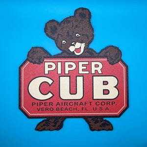 Peut inclure: Un panneau en forme d'ours brun tenant un panneau rouge et blanc sur lequel on peut lire "PIPER CUB". Le panneau comprend également le texte "PIPER AIRCRAFT CORP. VERO BEACH, FL. U.S.A." sur un fond bleu.