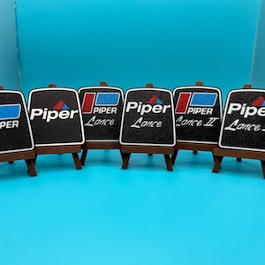 May include: Six small black signs with white text on easels. The signs read "Piper", "Piper Lance", "Piper Lance", "Piper Lance II", "Piper Lance II".