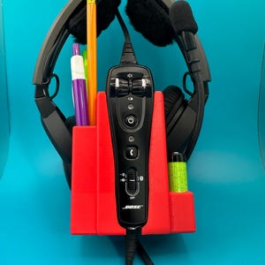 May include: A red plastic headset holder with a black Bose headset, a pencil, two pens, and a green battery. The holder has a built-in compartment for the headset and a separate compartment for the battery.