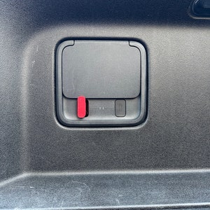 May include: A black plastic panel with a rectangular opening. The opening has a black cover with a red button on the left side. The cover is slightly ajar.