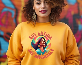 May include: A woman with curly brown hair is wearing a yellow sweatshirt with a colorful graphic design that says "Soy Latina Soy Imparable".
