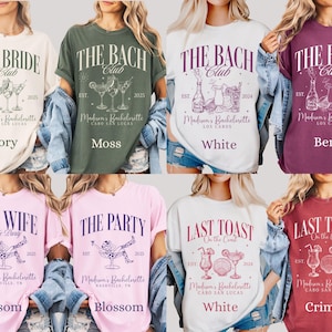 May include: Assorted-color graphic t-shirts with text like "The Bride Club" and "Last Toast on the Coast". Each shirt features a unique design with cocktail illustrations and the text "Madison's Bachelorette". Colors include ivory, moss, white, berry, blossom, and crimson.