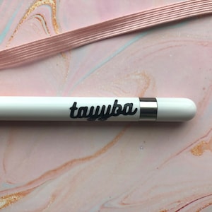 以下が含まれることがあります： 黒い筆記体で「tayyba」という名前が入った白いスタイラスペン。スタイラスペンの先端近くにはシルバーのバンドがあります。ピンクのゴムバンドが背景にあり、ピンクとゴールドの大理石模様があります。
