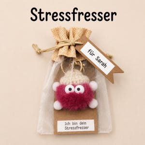 Könnte beinhalten: Ein Schlüsselanhänger in Form einer runden, flauschigen, dunkelrosa Kreatur mit weißen Akzenten und großen Augen, verpackt in einem transparenten Beutel. Ein braunes Papieretikett trägt "für Sarah" und der Aufdruck lautet "Ich bin dein Stressfresser".