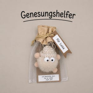 Könnte beinhalten: Eine beige gehäkelte Plüschfigur mit großen weißen Augen in einem kleinen transparenten Beutel mit Kordelzug. Ein braunes Papieretikett am Beutel trägt die Aufschrift "für Luisa" und ein weißes Etikett unten trägt "Ich beschütze dich, bis du wieder stark bist".