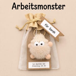 Könnte beinhalten: Ein beiger, flauschiger Monster-Schlüsselanhänger mit großen weißen Augen in einem transparenten Kordelzugbeutel. Ein braunes Etikett am Beutel trägt die Aufschrift "für Sarah" und ein weißes Etikett unten zeigt "Ich überlebe den Arbeitstag mit dir".
