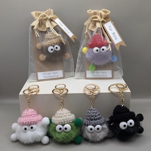 Könnte beinhalten: Handgemachte Monster-Schlüsselanhänger in Weiß, Grün, Grau und Schwarz. Jeder Schlüsselanhänger hat einen flauschigen Körper, große Kulleraugen und eine gehäkelte Mütze. Einige sind in Geschenkbeuteln mit Anhängern verpackt.