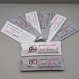 Könnte beinhalten: Eine Auswahl an Filz-Kartenhaltern in Grau und Weiß, mit dem Text "passt perfekt ins Team" in Rot und Schwarz. Die Halter haben einen Kartenschlitz und ein Smiley-Gesicht. Der Text "DANKE, FÜR DEINE WERTVOLLE ARBEIT!" ist ebenfalls zu sehen.